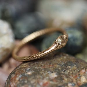May include: A gold-colored ring in the shape of a snake, with detailed scales and a textured surface. The snake's head is slightly raised, and the ring is resting on a collection of smooth, colorful stones.