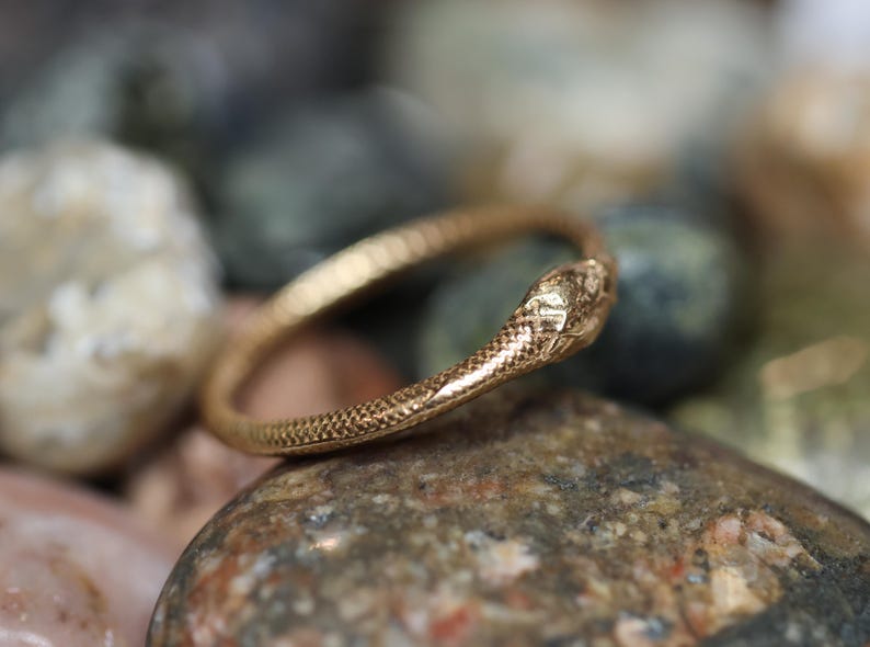 May include: A gold-colored ring in the shape of a snake, with detailed scales and a textured surface. The snake's head is slightly raised, and the ring is resting on a collection of smooth, colorful stones.