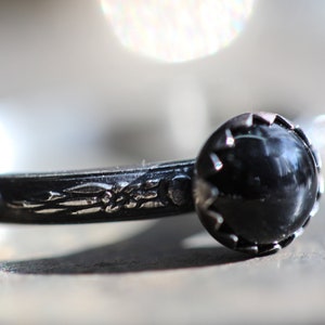Puede incluir: Un anillo de plata con una piedra preciosa negra engastada en un engaste de plata decorativo. La banda del anillo está adornada con un intrincado diseño floral.