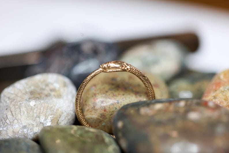 May include: A gold-colored ring in the shape of a snake, with detailed scales and a snake head. The ring is set against a backdrop of various smooth, colorful stones, creating a natural and textured contrast.