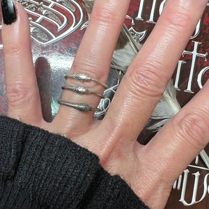 May include: Three silver snake rings are stacked on a finger. The rings have a detailed snake design. The background includes a book cover with ornate lettering and a feather. The person's nails are painted black.