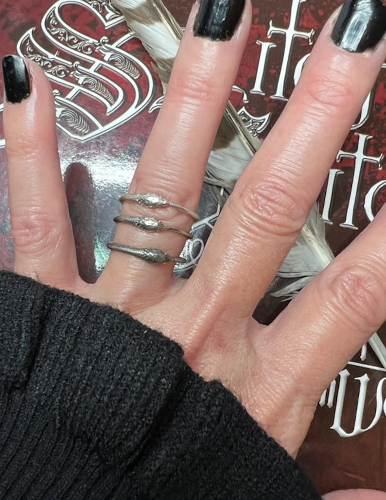 May include: Three silver snake rings are stacked on a finger. The rings have a detailed snake design. The background includes a book cover with ornate lettering and a feather. The person's nails are painted black.