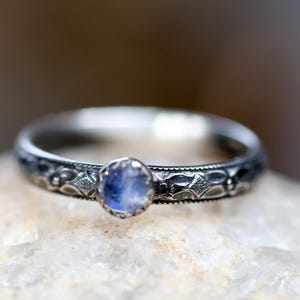 May include: A silver ring with a central oval, pale blue gemstone. The band is decorated with an engraved pattern. The ring is set against a blurred background, emphasising the intricate details of the jewellery.