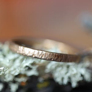 Puede incluir: Primer plano de un anillo de oro texturizado. El anillo tiene un acabado martillado y se encuentra sobre un fondo borroso de follaje blanco y verde. La superficie del anillo refleja la luz.
