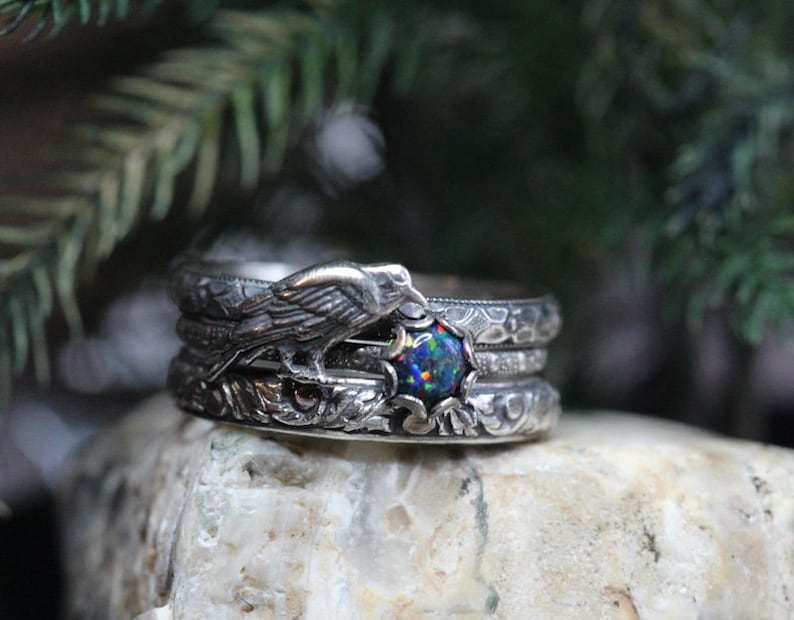 Puede incluir: Tres anillos de plata con un dise&ntilde;o intrincado. El anillo superior presenta un p&aacute;jaro posado en una rama con una piedra preciosa de &oacute;palo negro en el centro. Los anillos est&aacute;n sobre una roca gris.