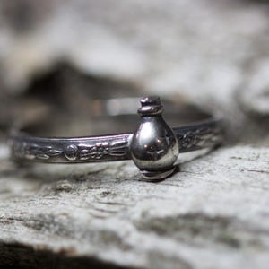 May include: A silver ring with a detailed design featuring a small bottle charm.