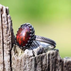 May include: A silver ring featuring an oval-shaped, dark red iridescent gemstone set in a scalloped metal bezel. The ring band has a textured, leaf-like pattern etched along its surface, providing a detailed finish to the metalwork.