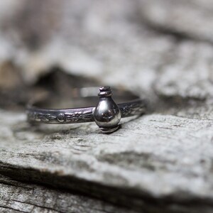 May include: A silver ring with a detailed design featuring a small, round bottle charm.