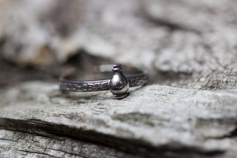 May include: A silver ring with a detailed design featuring a small, round bottle charm.