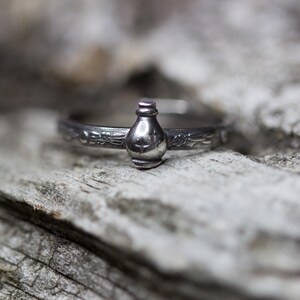 May include: A silver ring with a detailed design featuring a small bottle charm.