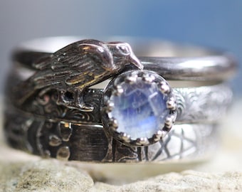 May include: A silver ring set with a moonstone and a bird design. The ring is made of three bands, each with a different texture.