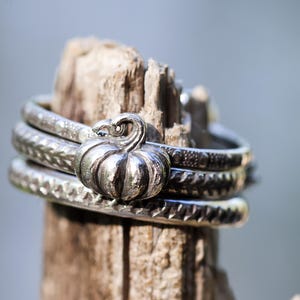May include: A set of three stacked silver rings featuring a central pumpkin charm. Each ring band displays a distinct textured pattern, including a braided design and a series of small circular indentations, all crafted in a polished silver metal.
