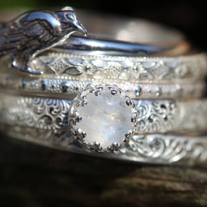 May include: A stack of five silver rings with intricate designs, featuring a moonstone ring with a silver setting in the center. A silver bird charm sits atop the rings.
