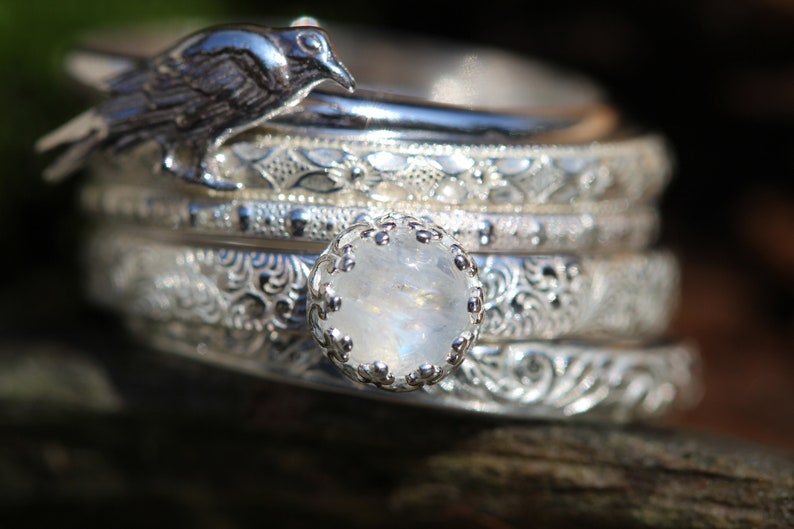 May include: A stack of five silver rings with intricate designs, featuring a moonstone ring with a silver setting in the center. A silver bird charm sits atop the rings.
