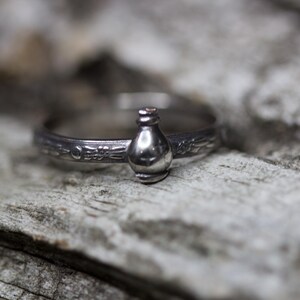 May include: A silver ring with a detailed design featuring a small bottle charm. The ring is on a rustic wooden surface.