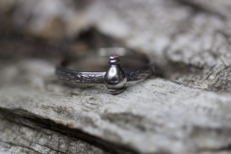 May include: A silver ring with a detailed design featuring a small bottle charm. The ring is on a rustic wooden surface.