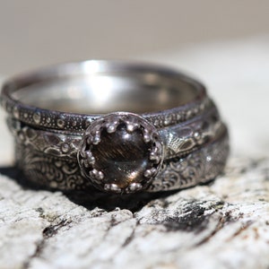 May include: A silver ring set with a dark brown gemstone. The ring is made up of four bands, each with a different design. The bands are stacked on top of each other.