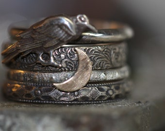May include: A set of four silver rings with intricate designs. The top ring features a silver raven with a crescent moon design. The rings are stacked on top of each other.