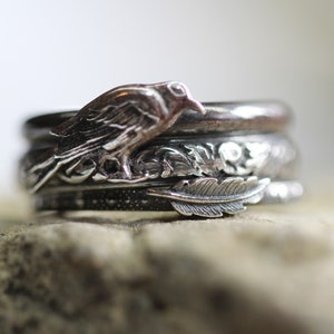 Puede incluir: Una pila de anillos de plata con diseños inspirados en la naturaleza. El anillo superior presenta un pájaro detallado, mientras que los anillos inferiores muestran motivos florales y de plumas. Los anillos se exhiben sobre una superficie texturizada.