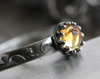 May include: A silver ring with a round, golden-yellow gemstone. The ring has a detailed floral and leaf pattern on the band and a crown-like setting around the stone. The ring is a piece of jewelry.
