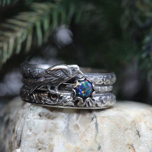 Puede incluir: Tres anillos de plata con un dise&ntilde;o intrincado. El anillo superior presenta un p&aacute;jaro posado en una rama con una piedra preciosa de &oacute;palo negro en el centro. Los anillos est&aacute;n sobre una roca gris.