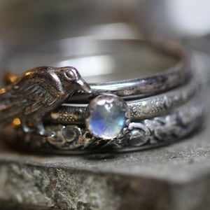 Puede incluir: Un anillo de plata con una piedra de luna y un peque&ntilde;o encanto de p&aacute;jaro. El anillo est&aacute; hecho de tres bandas, cada una con un dise&ntilde;o diferente.