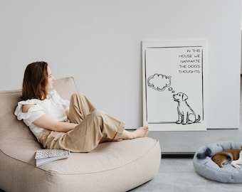May include: A woman sits on a light brown beanbag chair, looking to the right. A white canvas with black marker writing hangs on the wall behind her. The writing says "In this house we narrate the dog's thoughts". A black and white drawing of a dog with a thought bubble above its head is on the canvas. A small dog is lying in a gray and white dog bed on the floor.
