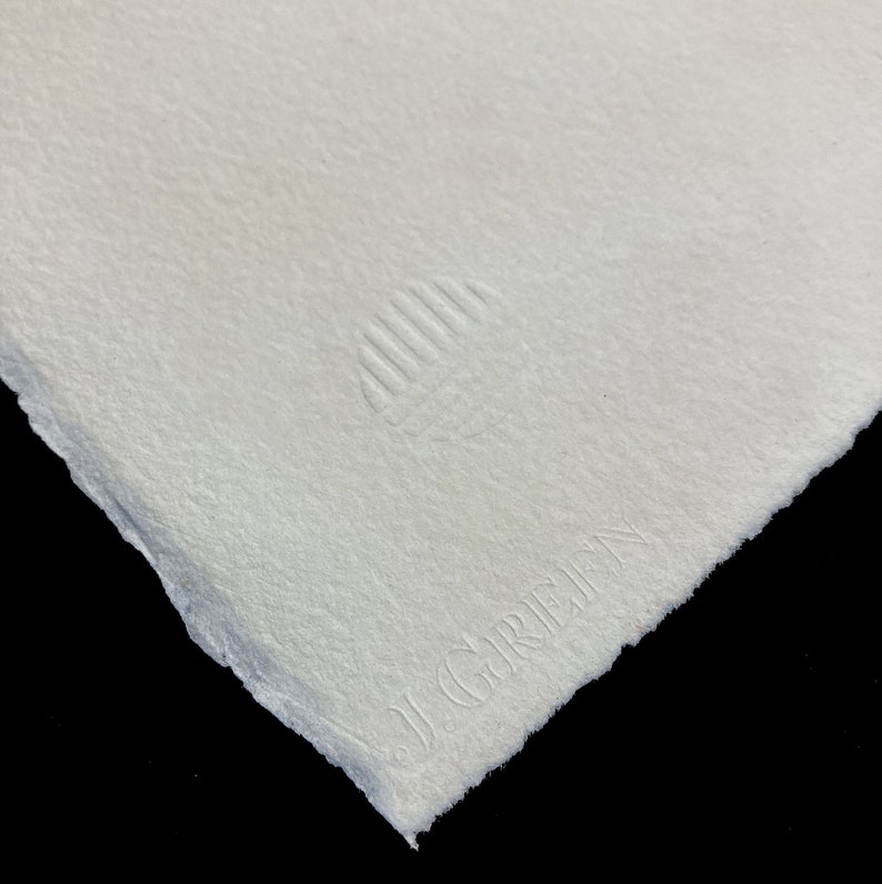 Puede incluir: Primer plano de un papel blanco con una superficie texturizada. El papel tiene un peque&ntilde;o logotipo en relieve que dice "J.G. Greif" en la esquina inferior derecha.