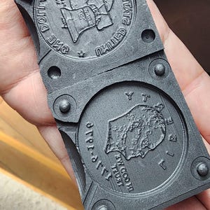Puede incluir: Dos moldes de plástico gris para fundir monedas. El molde superior tiene las palabras "UNITED STATES OF AMERICA" y "ONE DOLLAR" alrededor del borde. El molde inferior tiene las palabras "IN GOD WE TRUST" y "LIBERTY 1776" alrededor del borde.