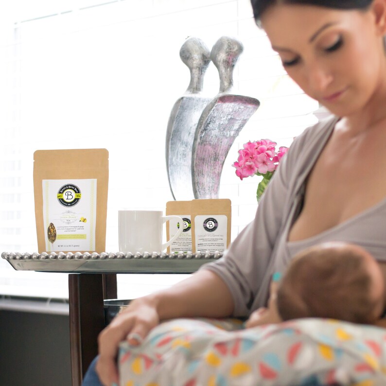 May include: A brown paper bag of tea with a white label and the text "Babe & Brew Tea" sits on a silver tray with a white mug and two smaller tea packets. A woman is breastfeeding a baby.