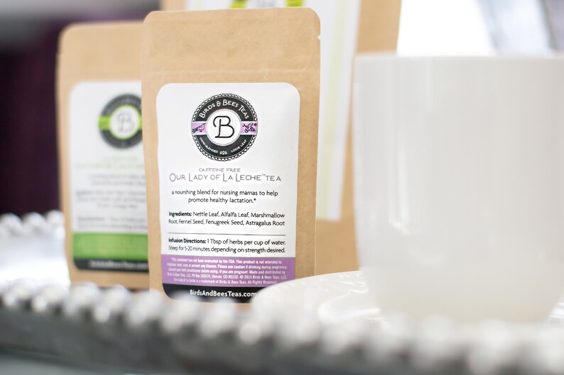 May include: Two brown paper tea pouches with white labels, featuring text and a logo. The text reads "Our Lady of La Leche Tea" and "Caffeine Free". A white teacup and saucer are in the foreground.