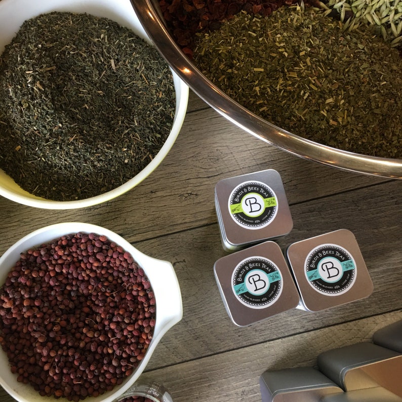 May include: Three silver tins of Bird & Bees Tea, each with a green and white label featuring the company logo and the text "Bird & Bees Tea". The tins are arranged on a wooden surface with other tea ingredients in bowls.