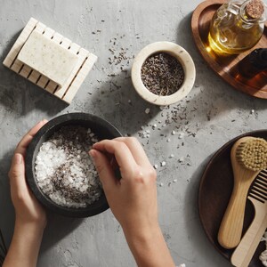 May include: A close-up of a person's hands adding dried lavender to a bowl of bath salts. The bowl is made of dark grey stone and is sitting on a light grey surface. There is a small bowl of dried lavender, a bar of soap, and a wooden tray with a glass bottle of oil in the background.