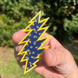 May include: A yellow lightning bolt sticker with a blue background and a pattern of yellow bicycles. The sticker is held in a hand.