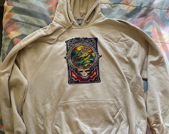 May include: A beige hooded sweatshirt with a graphic print of a colorful skull and a landscape scene. The skull is surrounded by a decorative frame.