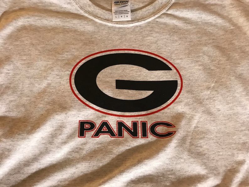 May include: A gray t-shirt with a black and red graphic of a large "G" inside a red oval with the word "PANIC" underneath.