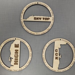 May include: Three round wooden ornaments with a laser-cut design of the Shawangunk Mountains in New York. The ornaments feature the text "SKY TOP", "HIGH E", and "CCK".