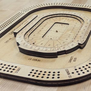 May include: A wooden game board shaped like a stadium with a capacity of 85,575. The board has a black border and a brown surface. The board has a start position and a finish position. The board is labeled with the street names "Henry St" and "Woodward Ave".