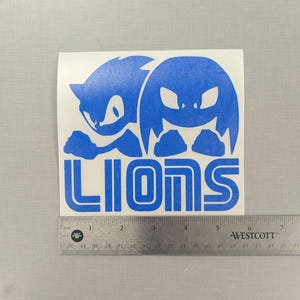May include: Blue vinyl decal with two cartoon characters, one with a blue spiky head and the other with a blue round head, both with white eyes. The characters are positioned above the word "LIONS" in blue.