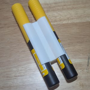 May include: Two yellow and black cylindrical objects with white labels and a white plastic connector. The objects are arranged side-by-side, connected by a white plastic piece.