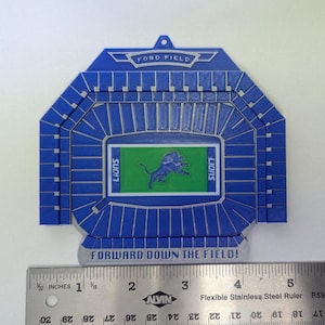 May include: A blue and white 3D-printed replica of Ford Field, home of the Detroit Lions. The stadium model features a green field with the Lions logo and the text "LIONS". The text "FORWARD DOWN THE FIELD!" is at the bottom.