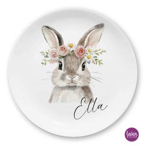 Könnte beinhalten: Weiße Keramikplatte mit einer Aquarellillustration eines Hasen mit Blumenkranz. Der Hase ist grau und braun mit einem süßen Ausdruck. Der Name "Ella" ist kursiv geschrieben. Die Platte hat ein "Levar Design"-Logo.