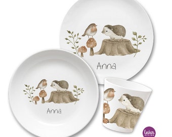 May include: A white children's dinnerware set with a woodland theme. The set includes a large plate, a bowl, and a cup. The design features a hedgehog, a robin, and mushrooms on a tree stump. The name "Anna" is written on each piece.