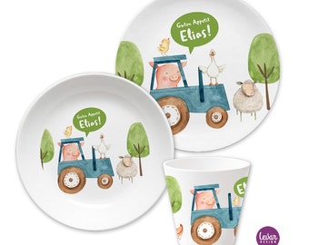 May include: A white children's dinnerware set with a blue tractor, a pink pig, a yellow chick, a white goose, and a white sheep. The set includes a large plate, a small plate, and a cup. The text "Guten Appetit Elias!" is on the plates.