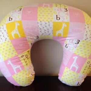 May include: A pink and yellow patchwork nursing pillow with a U-shape. The pillow features giraffe and number patterns. The pillow is designed for baby care and support.