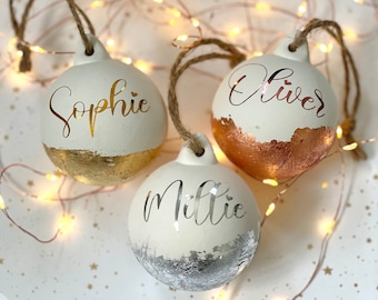 May include: Three white Christmas ornaments with names in gold, silver, and copper lettering. Each ornament has a metallic bottom edge and a twine loop for hanging. The names Sophie, Oliver, and Millie are displayed on the ornaments.