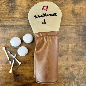 Könnte beinhalten: Ein beiger Golfschlägerkopfbezug mit braunem Lederboden. Der Bezug ist mit dem Namen "Weatherall" bestickt und trägt eine rote Flagge mit der Nummer "19". Drei weiße Golfbälle und drei Golfschläger liegen auf einer Holzoberfläche.