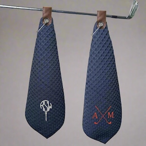 May include: Two blue waffle weave golf towels with leather hanging loops. One towel has the monogram "RSW" with a golf tee and the other has the monogram "AM" with crossed golf clubs.