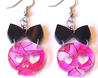 May include: Pink skull earrings with black bow accents. The skull has a cracked glass effect and heart-shaped eyes.