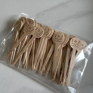 Pode incluir: Um saco de plástico transparente cheio de inúmeros palitos de madeira para bebidas. Cada palito tem as iniciais "I & J", a data "04.05.2024" e um símbolo de coração. Os palitos são feitos de madeira clara.